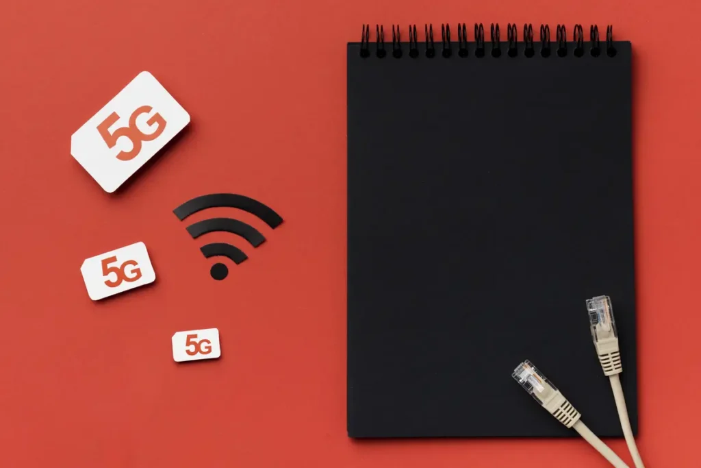 Top view of 5G SIM cards, Wi-Fi symbol, Ethernet cables, and a black spiral notebook on a red background — representing internet connectivity and travel tech setup